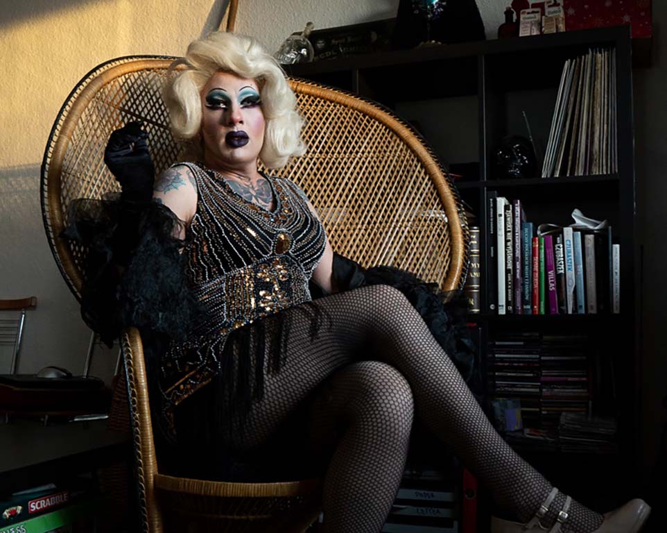 Image: Drag queen with blonde wig, blue eye makeup, black lips and long eyelashes, tattoo on the right shoulder, sitting in a wicker chair with right leg crossed over, black fishnet stockings, glitter bodice with fringes, a bookshelf in the background.