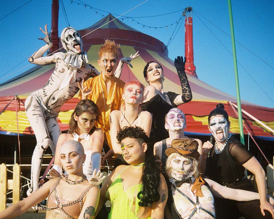 Bild: Venus Boys 10 junge Menschen vor einem Zirkuszelt aufgebaut alle in intensivem MakeUp und Kostümen in wilden Posen, teilweise die Zungen raustreckend.