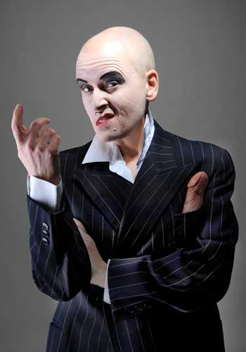 Image: Bridge Markland with shaved head, intense makeup and painted moustache, in pinstriped suit with penis in the chest pocket, head turned to the left, grinning mischievously, the right arm bent, index finger loosely pointing upwards.