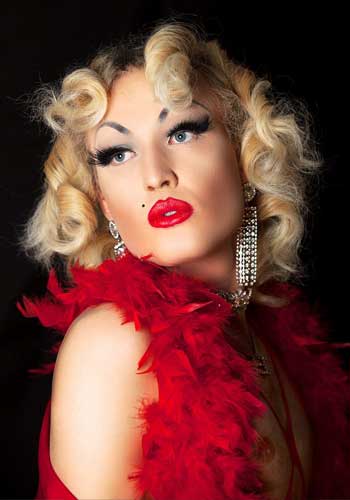 Bild: Olive Baldwin blickt über ihre rechte Schulter, mit intensivem Make-Up, roten schmollenden Lippen, langen Glitzerohrringen, lockigem blondem Haar und einer roten Federboa.
