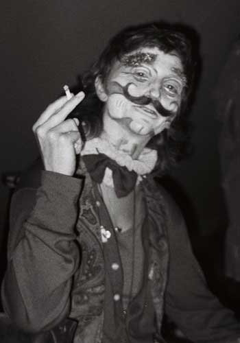 Image: Alec M. Balls in grotesque clownish make-up with large painted mustache, right arm bent with cigarette in hand.